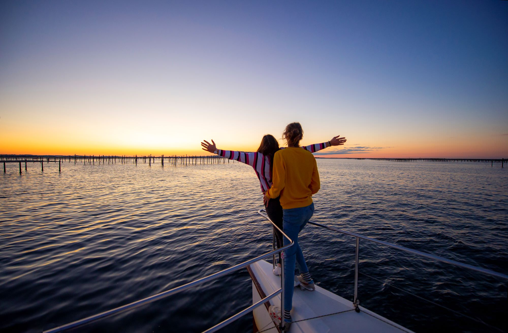 Un couple de jeunes en train de regarder le coucher du soleil sur l'étang de Thau sur un bateau-catamaran, devant les parcs à huîtres et Sète en toile de fond