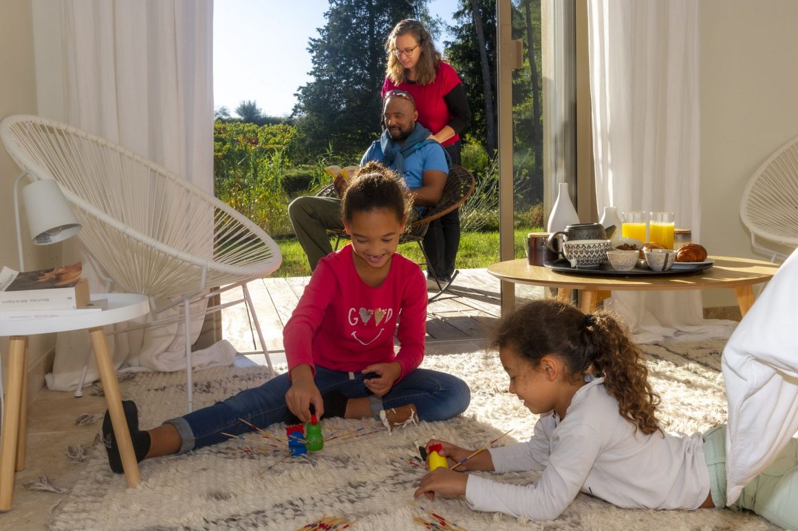 Une famille dans une chambre, avec deux enfants qui jouent et les parents qui lisent un livre sur la terrasse ensoleillée, avec en toile de fond les vignes de l'Hérault