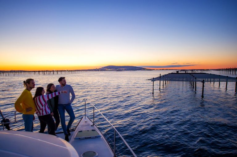 Un groupe de jeunes sur un bateau-catamaran en train de regarder le coucher du soleil sur l'étang de Thau, devant les parcs à huîtres et Sète en toile de fond