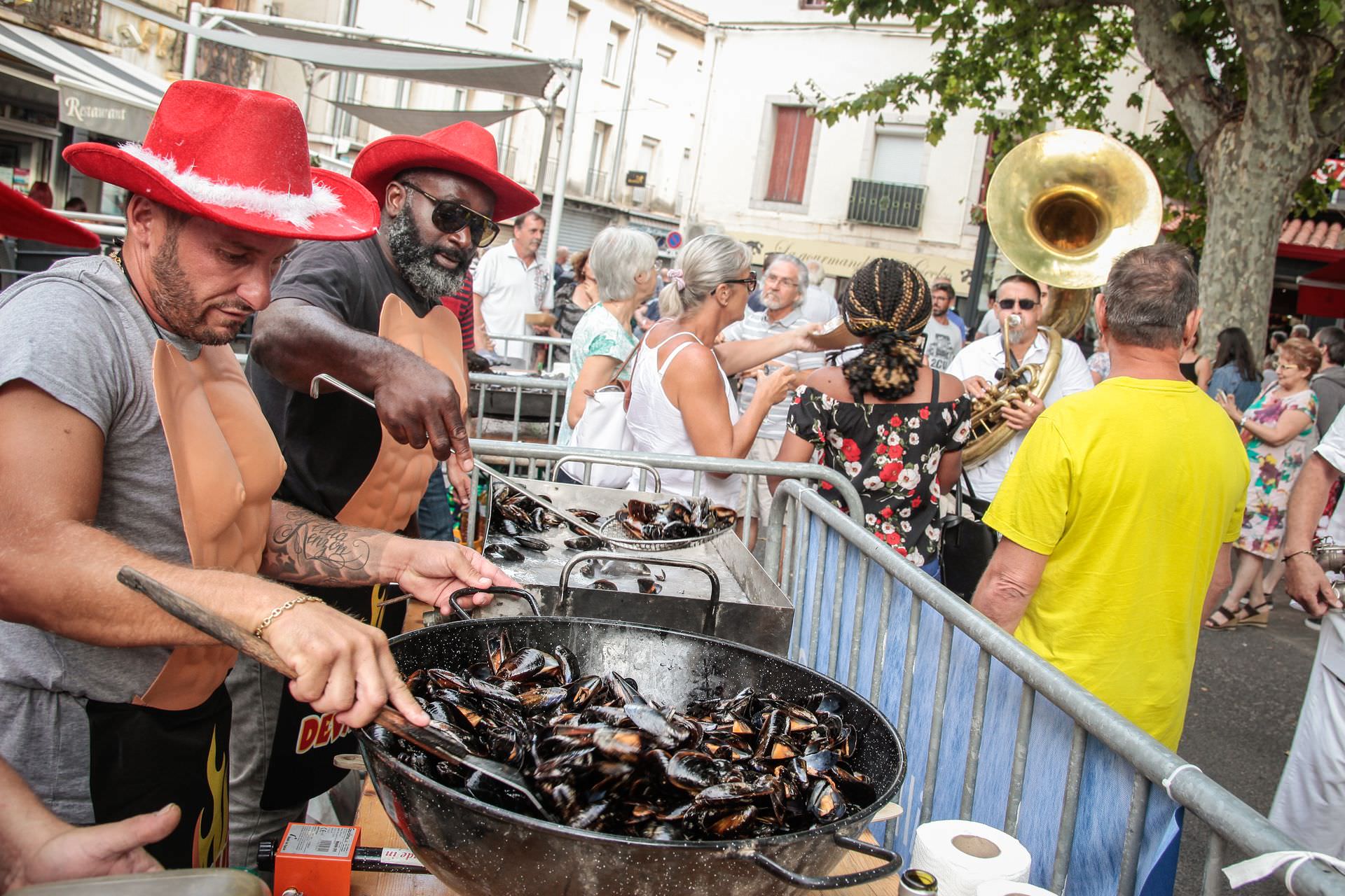 2 hommes qui cuisinent des moules dans une grande marmite lors du Festival de brasucade de Moules Total Musclum à Frontignan avec en arrière-plan une fanfare et des personnes qui profitent de l'ambiance