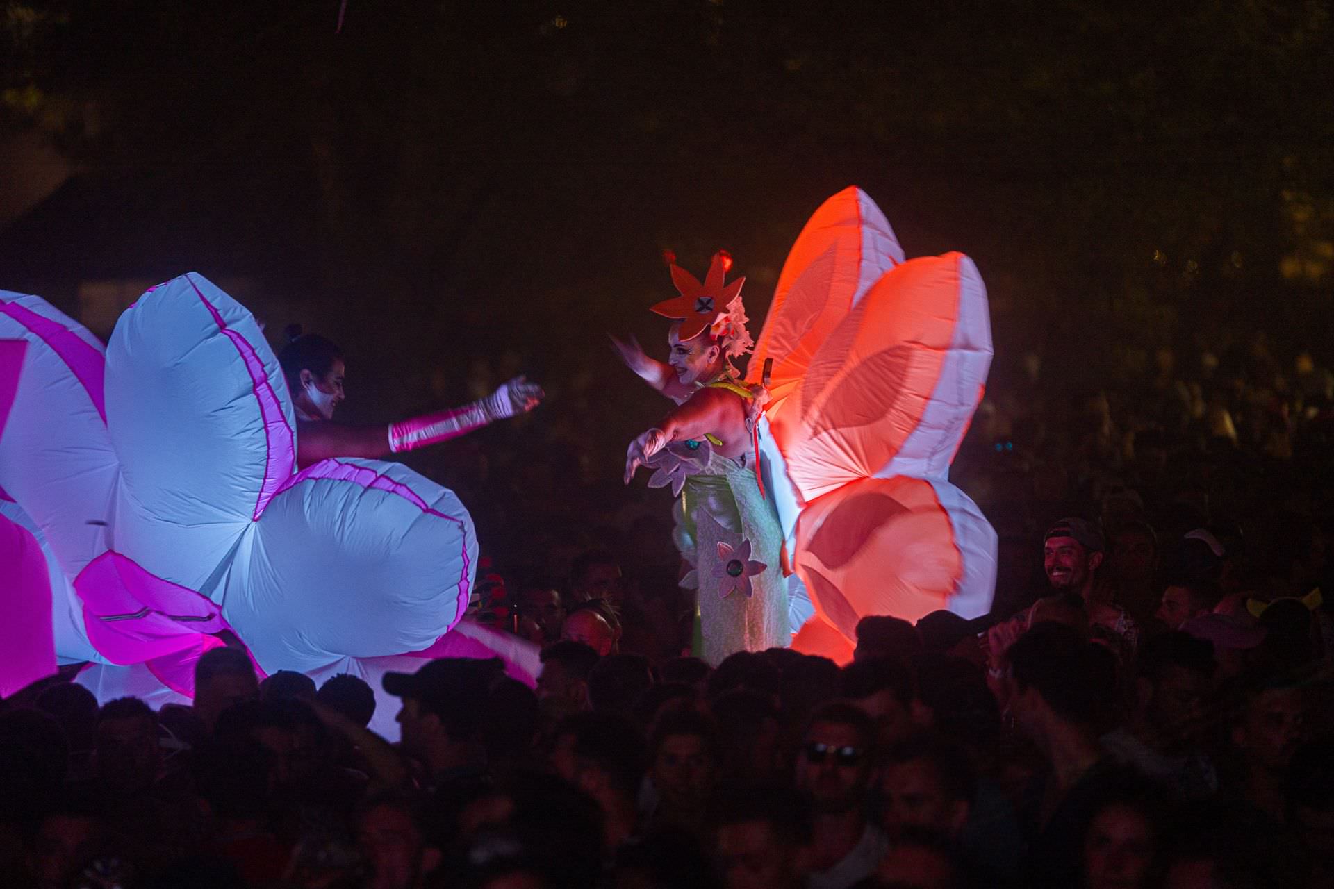 Personnes déguisées en fleurs fluorescentes sur échasses au milieu de festivaliers qui dansent à la nuit tombée lors de la Family Piknik au Domaine de Grandmont à Montpellier