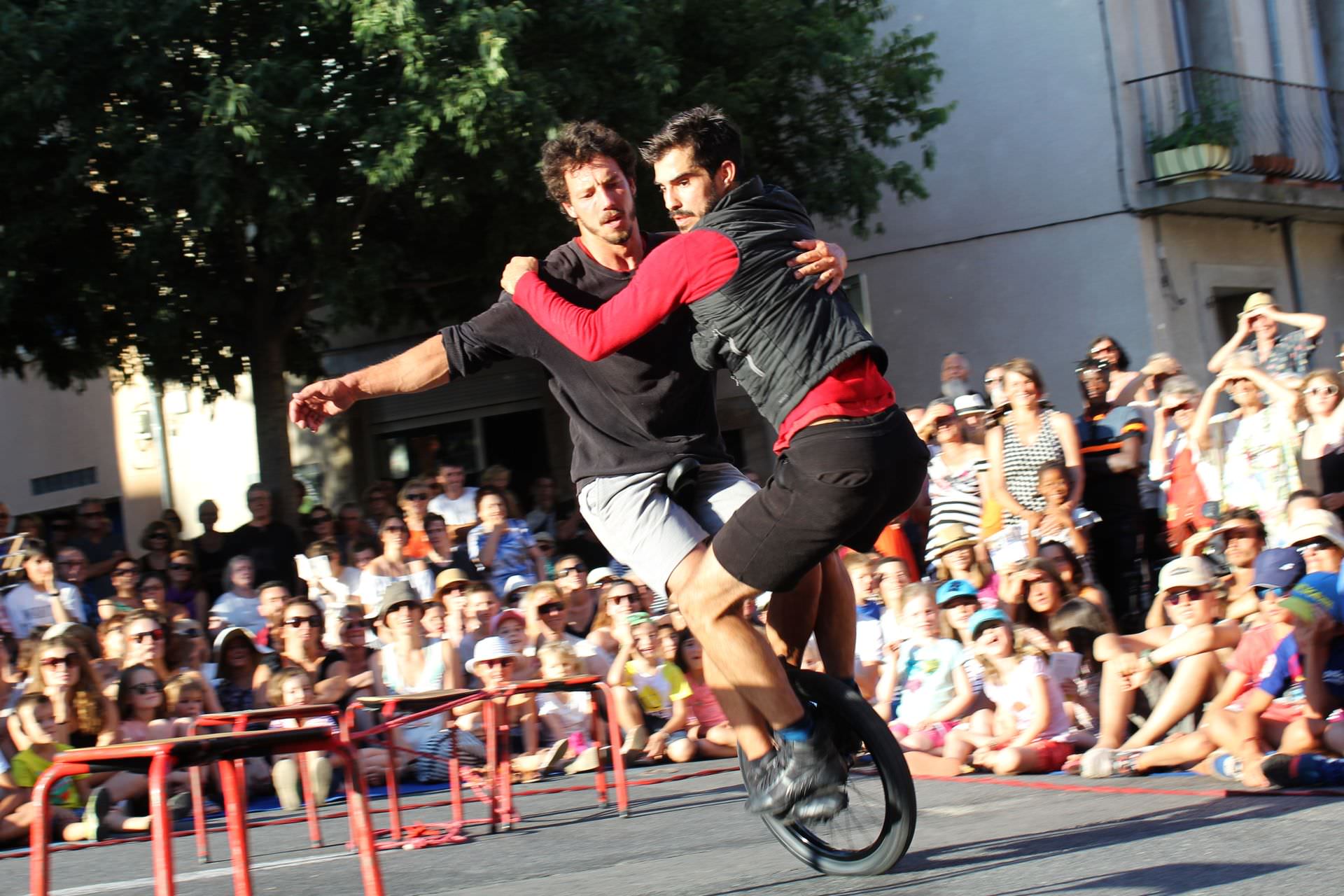 Duo d'acrobates du cirque Bankal en train de pédaler sur une même roue devant des spectateurs en famille sur une place de Lodève lors du Festival Résurgence en été