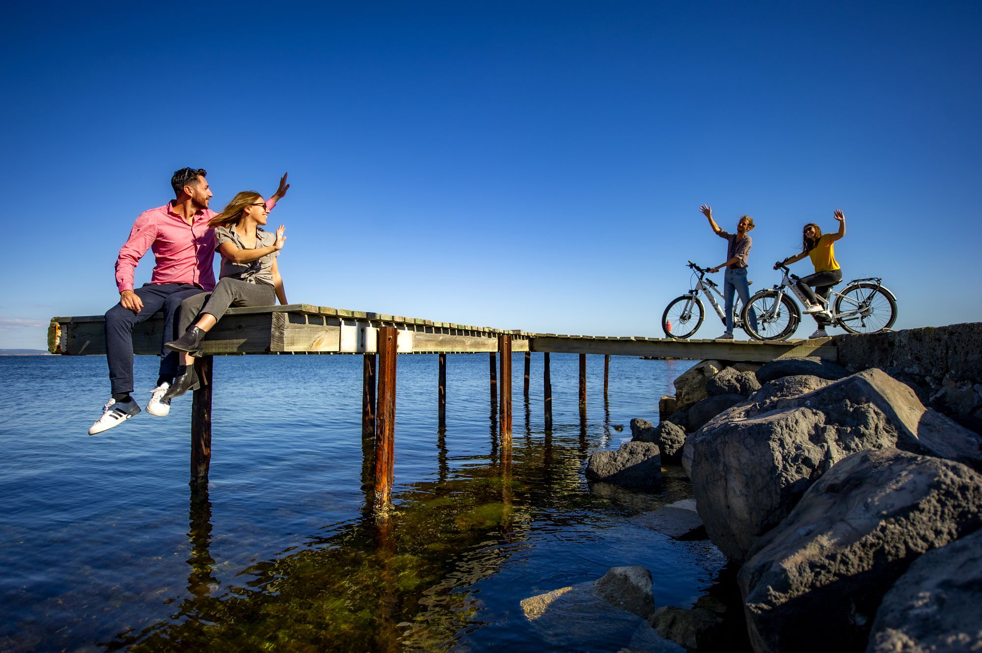 Couple de jeunes assis sur un ponton au dessus de l'étang de Thau, faisant signe à un couple de jeunes arrivant en vélo