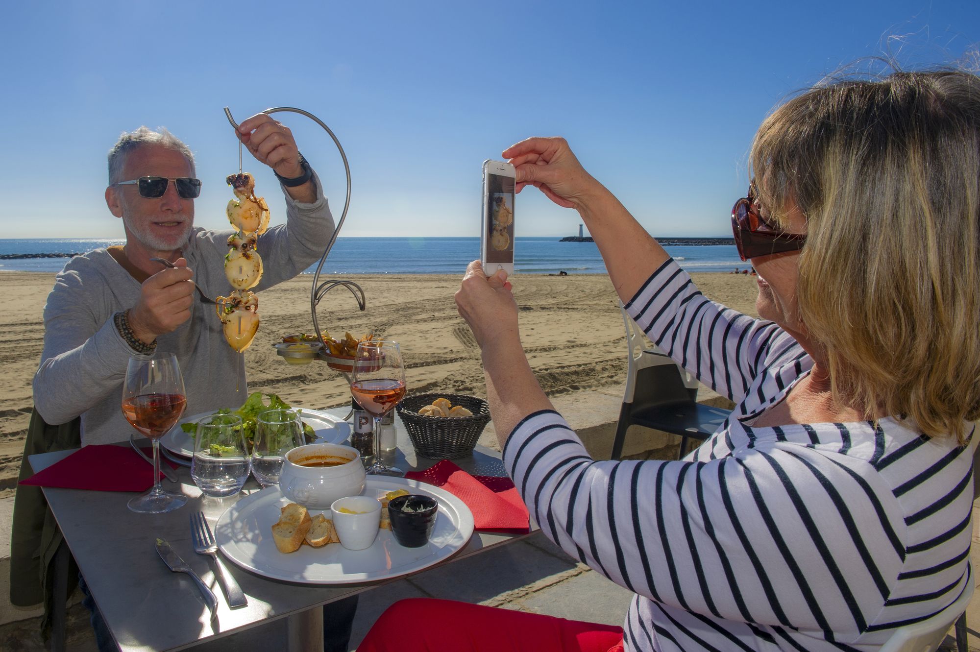 couple de jeuniors, quincados, en train de manger au restaurant, et de se prendre en photo avec la plage et la mer juste derrière eux au Cap d'Agde