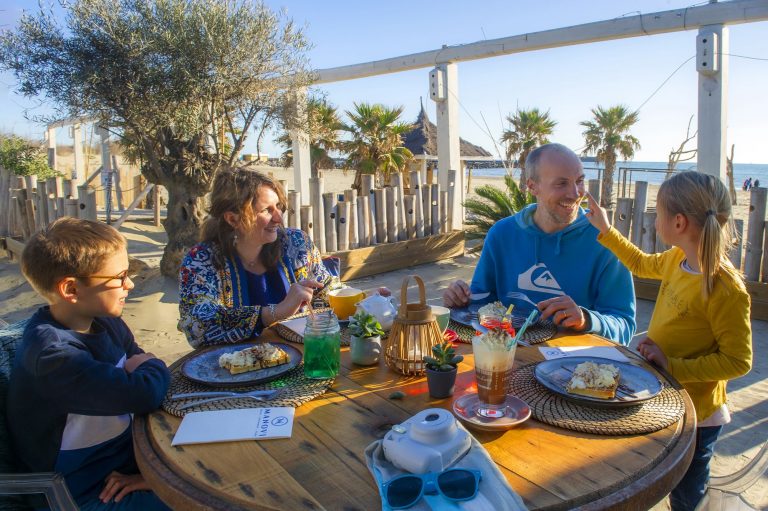 Famille avec deux enfants, en train de manger au restaurant, en train de rigoler, avec la plage et la mer juste derrière eux au Cap d'Agde