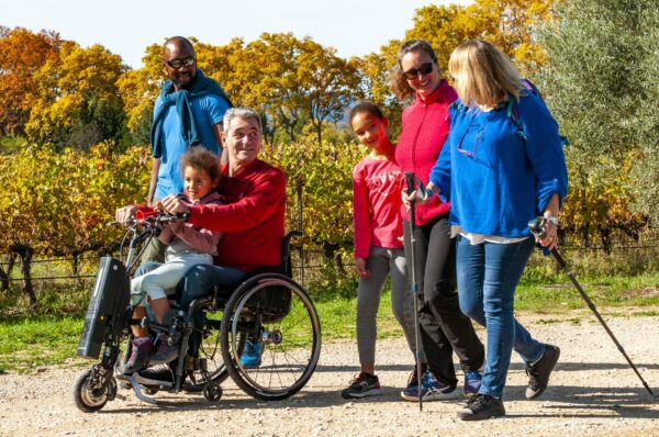 Une famille, enfants, petits enfants et grands parents avec le grand-père en fauteuil roulant, en train de se balader sur un chemin au coeur des vignes de l'Hérault