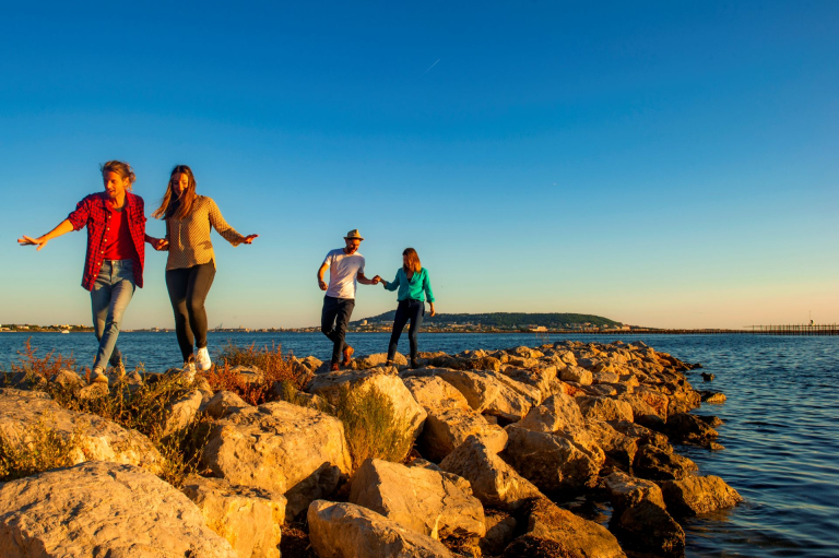 4 jeunes gens marchent allégrement sur la jetée de Bouzigues face à Sète lors du début du coucher de soleil. Des couleurs bleues, jaune oranger éclairent le paysage