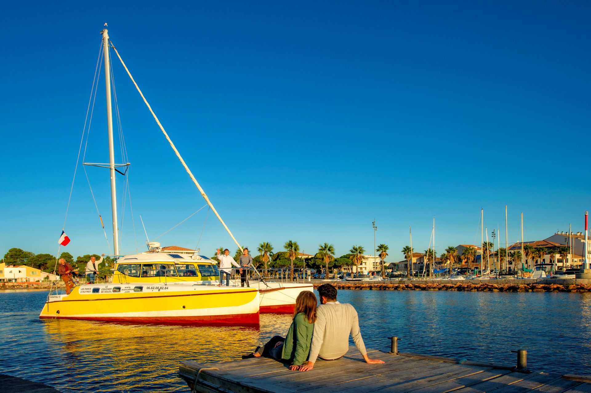 2 personnes assises sur un ponton de pêche regardent passer le catamaran Mansathau qui sort du port de Mèze. Il fait beau