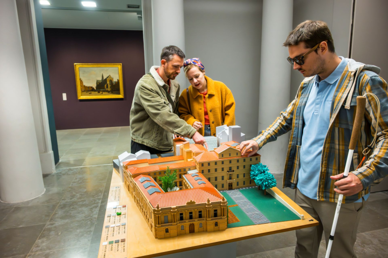 Une personne en situation de handicap visuel, touche la maquette du musée Fabre accompagné par 2 amis qui testent avec lui