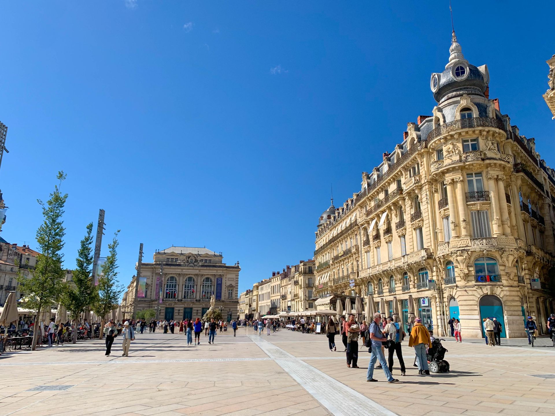 La place de la Comédie est sous un beau ciel bleu. Au bout de la place, se trouve le théâtre de la Comédie, en premier plan un bâtiment haussmannien imposant